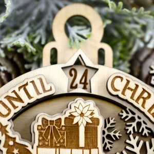 Puede incluir: Un adorno de madera para la cuenta atr&aacute;s de Navidad con una ventana en forma de estrella que muestra el n&uacute;mero 24. El adorno dice "Hasta Navidad" y tiene un dise&ntilde;o festivo con copos de nieve y una caja de regalo.