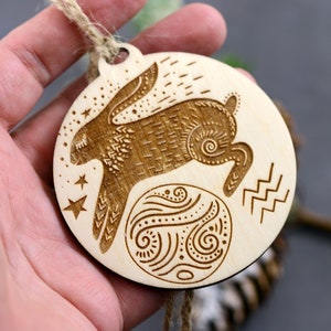 May include: A wooden ornament with a laser-cut design of a rabbit jumping over a circle with a swirling pattern. The ornament is hanging from a twine string and has the astrological symbol for Aquarius.