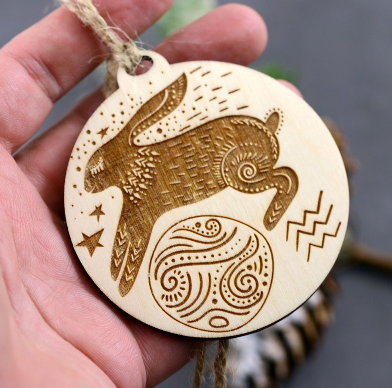 May include: A wooden ornament with a laser-cut design of a rabbit jumping over a circle with a swirling pattern. The ornament is hanging from a twine string and has the astrological symbol for Aquarius.