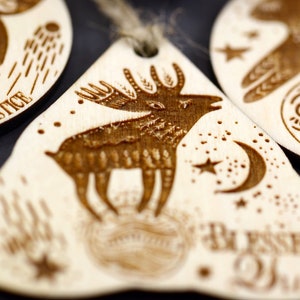 May include: A wooden ornament with a laser-etched design of a deer standing on a moon with stars. The ornament is light brown and has the text "Be Present" on the bottom.