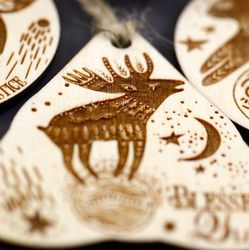May include: A wooden ornament with a laser-etched design of a deer standing on a moon with stars. The ornament is light brown and has the text "Be Present" on the bottom.