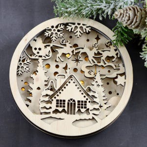Pu&ograve; includere: Un ornamento natalizio in legno con un design a taglio di una casa, alberi e la slitta di Babbo Natale con le renne. L'ornamento &egrave; illuminato da luci bianche calde.