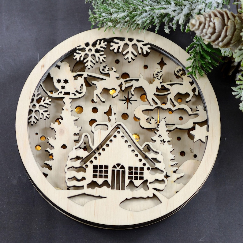 Pu&ograve; includere: Un ornamento natalizio in legno con un design a taglio di una casa, alberi e la slitta di Babbo Natale con le renne. L'ornamento &egrave; illuminato da luci bianche calde.