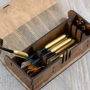 May include: A wooden box with a hinged lid, containing a set of six darts with black shafts and gold flights. The box has a laser-etched design on the lid.