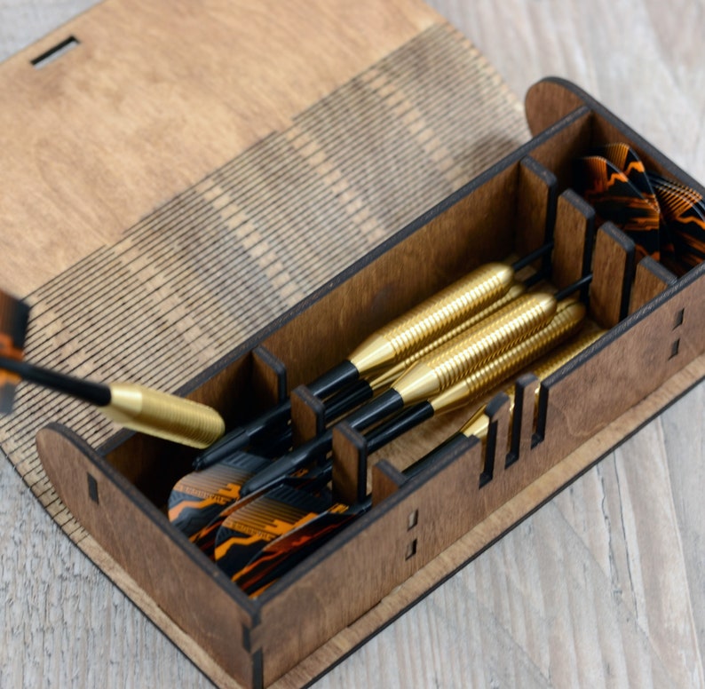 May include: A wooden box with a hinged lid, containing a set of six darts with black shafts and gold flights. The box has a laser-etched design on the lid.