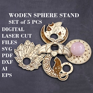 May include: Set of five laser-cut wooden sphere stands with various designs, including a crescent moon with a cat, a moth, crystals, and a crescent moon with mushrooms. The stands are designed to hold a sphere, and one stand has a pink sphere in place.