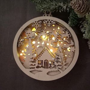 Pu&ograve; includere: Un ornamento natalizio in legno con un design illuminato. L'ornamento presenta una casa, alberi e una slitta di Babbo Natale con renne. L'ornamento &egrave; illuminato da luci LED bianco caldo.