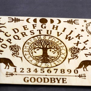 K&ouml;nnte beinhalten: Ein Ouija-Brett aus Holz mit einem Lebensbaum-Design in der Mitte. Das Brett enth&auml;lt das Alphabet, die Zahlen 1-9 und 0 sowie die W&ouml;rter "GOODBYE" und "YES".