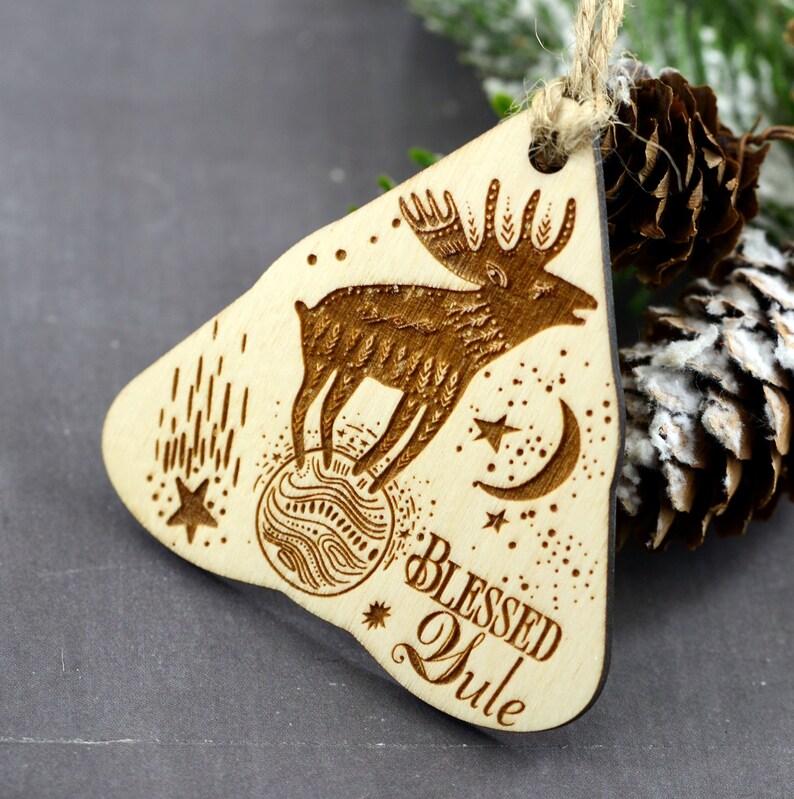 May include: Wooden Christmas ornament with a laser-engraved design of a moose standing on a sphere with stars and a crescent moon. The ornament is light brown and has the text "Blessed Yule" engraved on the bottom.
