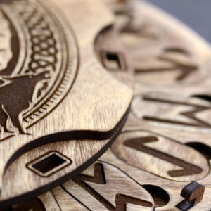 Puede incluir: Primer plano de un rompecabezas circular de madera con intrincados tallados. La pieza superior presenta un dise&ntilde;o de nudo celta y una figura de animal estilizada. Las piezas inferiores est&aacute;n superpuestas y muestran varios s&iacute;mbolos y patrones.