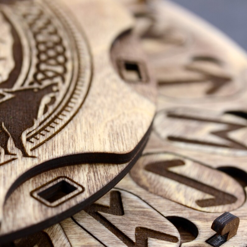 Puede incluir: Primer plano de un rompecabezas circular de madera con intrincados tallados. La pieza superior presenta un dise&ntilde;o de nudo celta y una figura de animal estilizada. Las piezas inferiores est&aacute;n superpuestas y muestran varios s&iacute;mbolos y patrones.