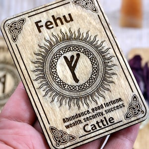 May include: Wooden rune card with the symbol for Fehu, the first rune in the Elder Futhark. The card is decorated with a sun and a Celtic knot. The text on the card reads "Fehu, Abundance, good fortune, wealth, security, success, Cattle".