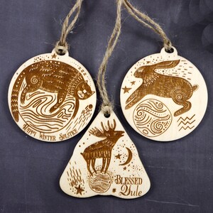 May include: Three wooden ornaments with a winter solstice theme. The first ornament is round and features a cat sleeping on a wave with the text "Happy Winter Solstice". The second ornament is round and features a rabbit jumping over a moon with stars. The third ornament is shaped like a bell and features a moose standing on a moon with the text "Blessed Yule".