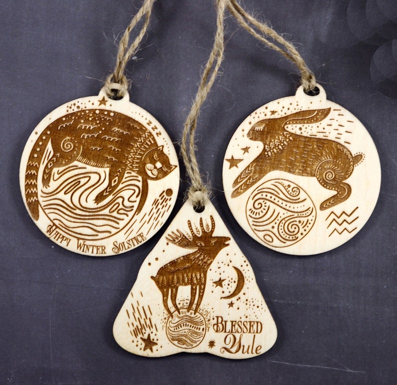 May include: Three wooden ornaments with a winter solstice theme. The first ornament is round and features a cat sleeping on a wave with the text "Happy Winter Solstice". The second ornament is round and features a rabbit jumping over a moon with stars. The third ornament is shaped like a bell and features a moose standing on a moon with the text "Blessed Yule".