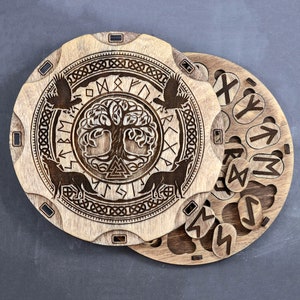 Puede incluir: Una caja de madera con una tapa circular que presenta un dise&ntilde;o tallado del &aacute;rbol de la vida Yggdrasil rodeado de runas. La tapa se abre para revelar un conjunto de runas de madera.