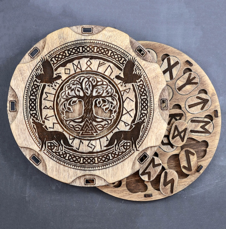Puede incluir: Una caja de madera con una tapa circular que presenta un dise&ntilde;o tallado del &aacute;rbol de la vida Yggdrasil rodeado de runas. La tapa se abre para revelar un conjunto de runas de madera.