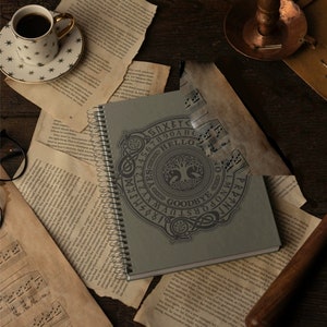 K&ouml;nnte beinhalten: Ein graues Spiralnotizbuch mit einem schwarz-wei&szlig;en Ouija-Brett-Design auf dem Cover. Das Design zeigt einen Lebensbaum mit den Worten "Goodbye" und "Hello" auf dem Brett.