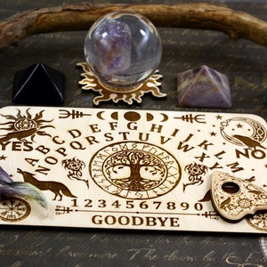 K&ouml;nnte beinhalten: Holz-Ouija-Brett mit einem Lebensbaum-Design, Alphabet, Zahlen, Ja, Nein, Auf Wiedersehen und einem Planchette. Das Brett ist von Kristallen und einer Kristallkugel umgeben.