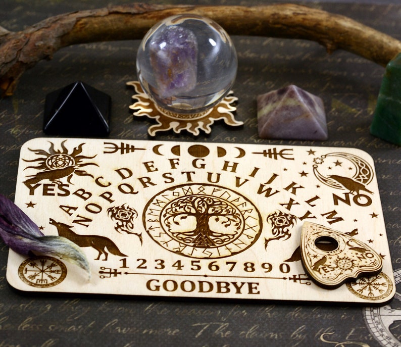 K&ouml;nnte beinhalten: Holz-Ouija-Brett mit einem Lebensbaum-Design, Alphabet, Zahlen, Ja, Nein, Auf Wiedersehen und einem Planchette. Das Brett ist von Kristallen und einer Kristallkugel umgeben.