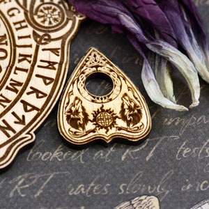 K&ouml;nnte beinhalten: Eine Holzplanchette mit einem Sonnen- und zwei Wolfsmotiven. Die Planchette hat eine dreieckige Form mit einem Loch in der Mitte. Die Planchette liegt auf einer dunkelgrauen Oberfl&auml;che mit Schrift.