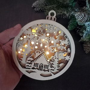 Pu&ograve; includere: Un ornamento natalizio in legno a forma di sfera con un design a taglio di una scena invernale. L'ornamento &egrave; illuminato da luci LED bianco caldo, creando un effetto luminoso. La scena include una casa, alberi, fiocchi di neve e una renna che tira una slitta.