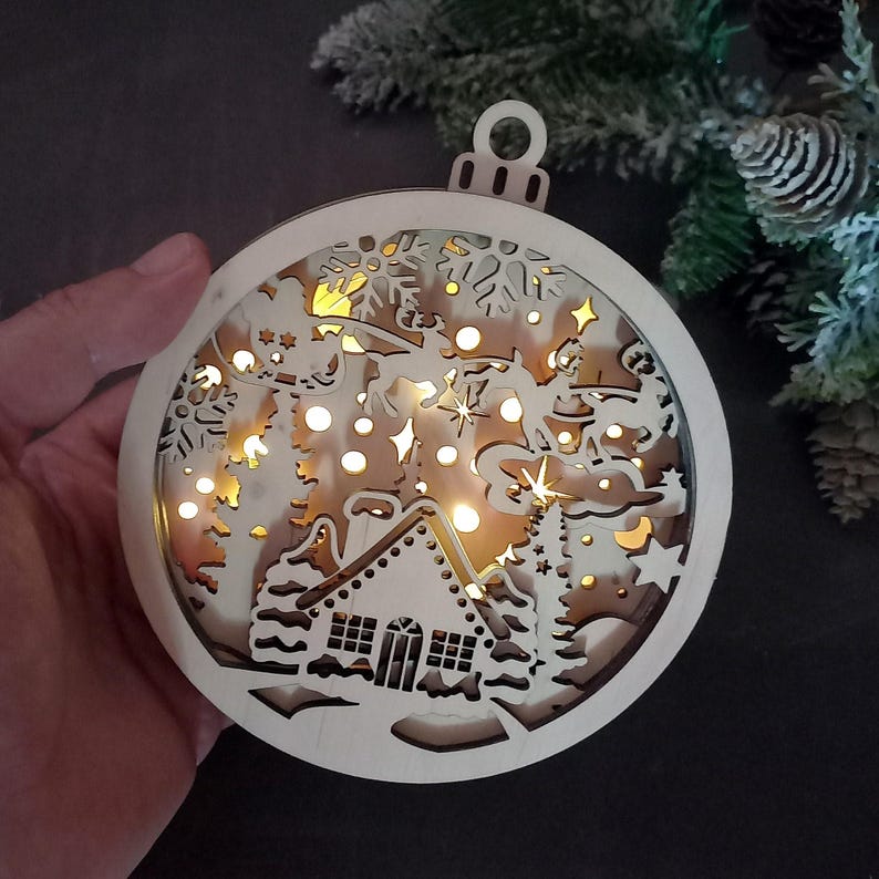 Pu&ograve; includere: Un ornamento natalizio in legno a forma di sfera con un design a taglio di una scena invernale. L'ornamento &egrave; illuminato da luci LED bianco caldo, creando un effetto luminoso. La scena include una casa, alberi, fiocchi di neve e una renna che tira una slitta.