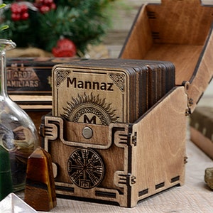 May include: Wooden box with a hinged lid, containing a set of wooden cards with the word "Mannaz" engraved on the top card. The box is decorated with a Celtic knot design and a compass.