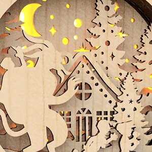 May include: A wooden Christmas ornament with a cutout design of a house, a tree, a crescent moon, and a figure. The ornament is illuminated with warm white LED lights.