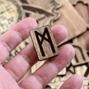 May include: A wooden rune with the symbol "M" carved into it. The rune is brown and has a natural wood grain. It is being held in a hand.