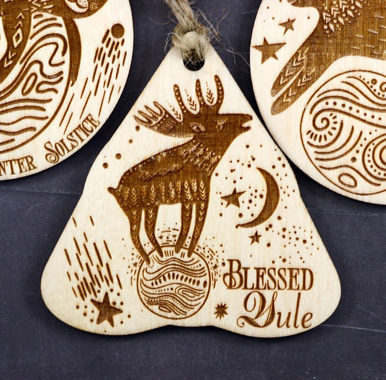 May include: A wooden ornament with a brown reindeer standing on a globe with stars and a crescent moon. The ornament is inscribed with the text "Blessed Yule".