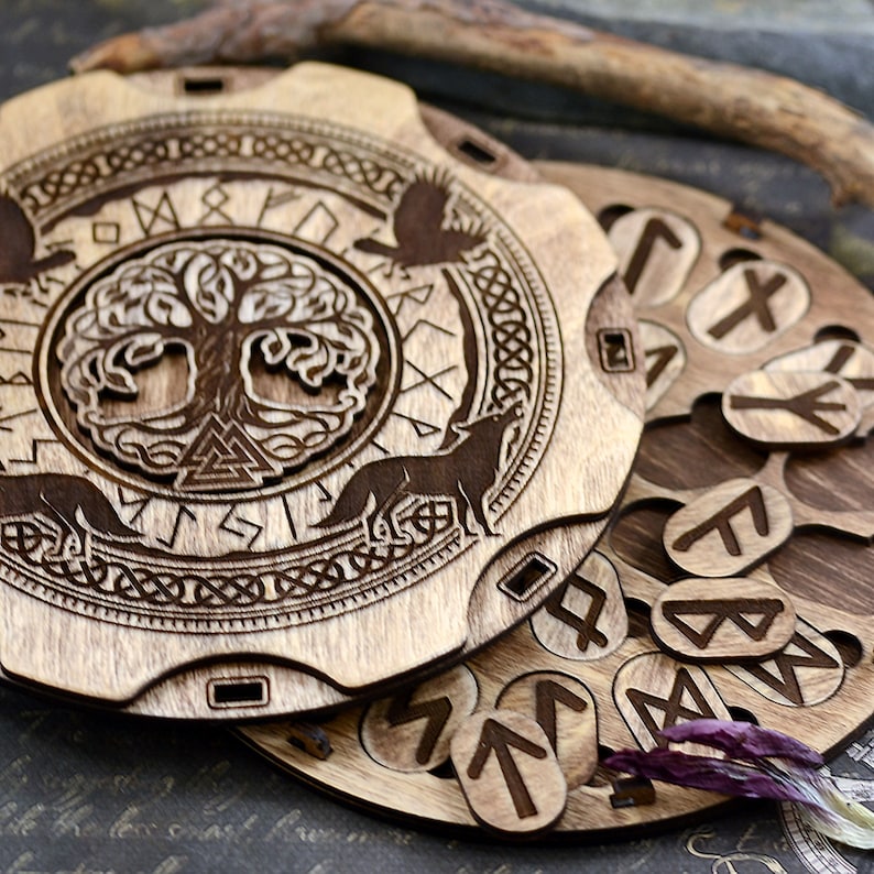 Puede incluir: Un tablero circular de madera con un &aacute;rbol de la vida Yggdrasil tallado rodeado de runas y lobos. El tablero tiene una ranura para 24 runas de madera individuales, cada una con una runa diferente tallada en ella.