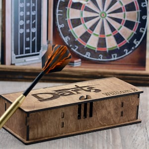 May include: A wooden box with the word "Darts" engraved on the top. The box has a slot for darts and the name "William B" is engraved on the side. A dart with orange and black feathers is in the foreground.