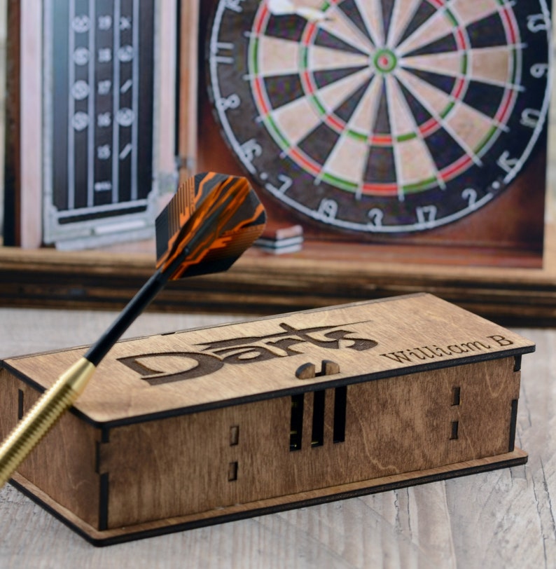 May include: A wooden box with the word "Darts" engraved on the top. The box has a slot for darts and the name "William B" is engraved on the side. A dart with orange and black feathers is in the foreground.