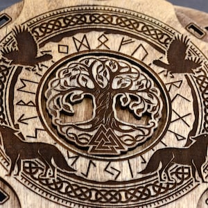 Puede incluir: Una placa redonda de madera con un dise&ntilde;o de &aacute;rbol de la vida tallado rodeado de un c&iacute;rculo de runas n&oacute;rdicas. Dos lobos est&aacute;n tallados a cada lado del &aacute;rbol de la vida.