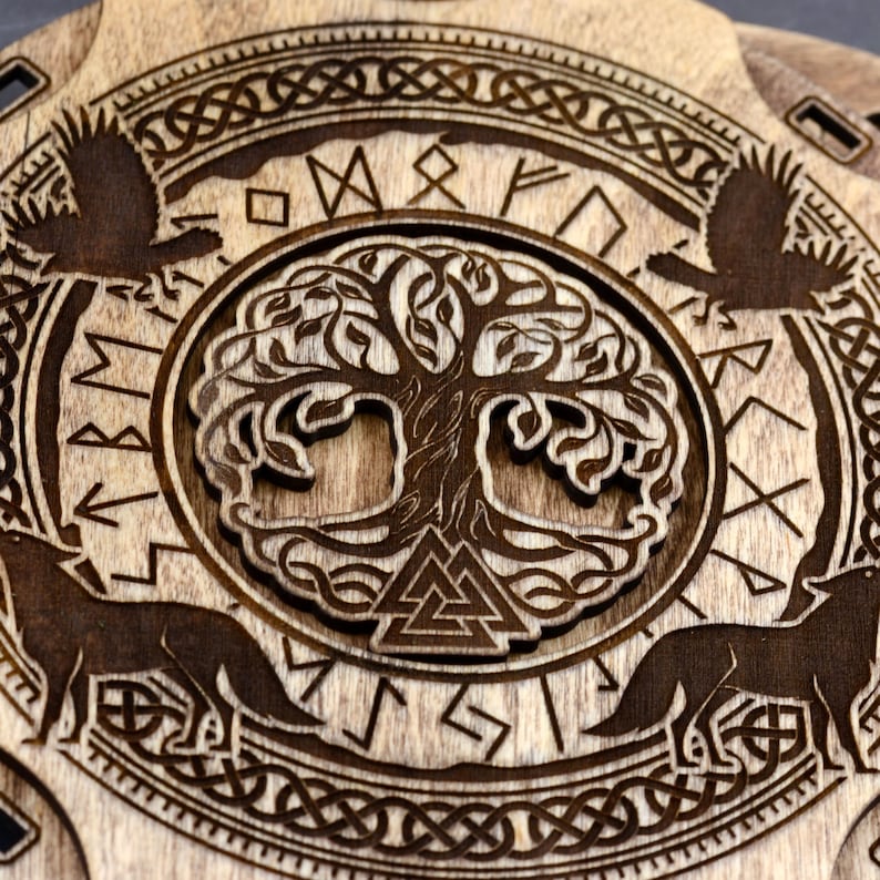 Puede incluir: Una placa redonda de madera con un dise&ntilde;o de &aacute;rbol de la vida tallado rodeado de un c&iacute;rculo de runas n&oacute;rdicas. Dos lobos est&aacute;n tallados a cada lado del &aacute;rbol de la vida.