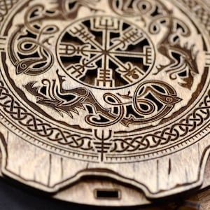 K&ouml;nnte beinhalten: Eine runde Holzplakette mit einer detaillierten Schnitzerei einer Kompassrose, die von zwei Drachen umgeben ist. Die Kompassrose ist ein traditionelles Symbol f&uuml;r Richtung und F&uuml;hrung. Die Drachen sind ein Symbol f&uuml;r St&auml;rke und Macht. Die Plakette ist aus hellbraunem Holz gefertigt und hat eine nat&uuml;rliche Oberfl&auml;che.