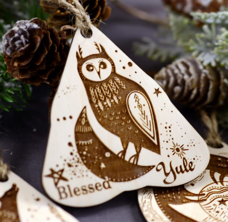 May include: Wooden Christmas ornament with a laser-etched design of an owl, moon, and stars. The ornament is painted brown and has the words "Blessed" and "Yule" inscribed on it.