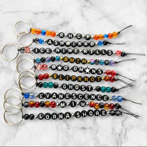 May include: A collection of ten keychains featuring silver metal rings and black letter beads spelling out band names including RHCP, Ashnikko, Ice Nine Kills, BMTH, Bad Omens, Slipknot, Ghost, Evanescence, MIW, and Lorna Shore, accented with colourful beads.