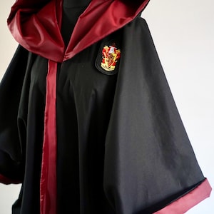 Puede incluir: Toga negra con capucha y ribete burdeos, con un parche del escudo de Gryffindor. La toga tiene mangas anchas y un acabado satinado, ideal para cosplay o eventos temáticos.