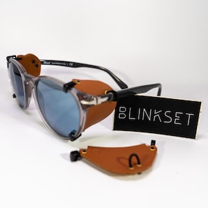 May include: Clear framed sunglasses with brown leather side shields and a black Blinkset logo on a black card.