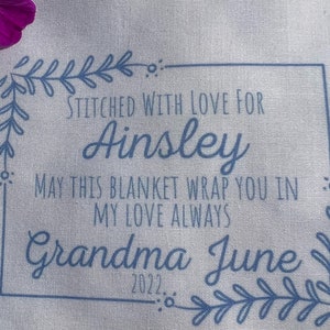 May include: A white fabric with blue embroidery that reads "Stitched with love for Ainsley. May this blanket wrap you in my love always. Grandma June 2022."