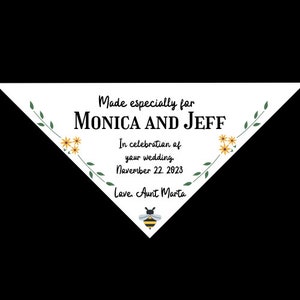 Peut inclure: Une faveur de mariage triangulaire blanche avec des feuilles vertes et des fleurs jaunes. Le texte indique "Made especially for Monica and Jeff. In celebration of your wedding. November 22, 2023. Love, Aunt Marta". Une abeille de dessin animé se trouve en bas.