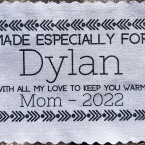 May include: White fabric tag with black text that reads "MADE ESPECIALLY FOR Dylan WITH ALL MY LOVE TO KEEP YOU WARM Mom - 2022". The tag is decorated with a black and white chevron pattern.