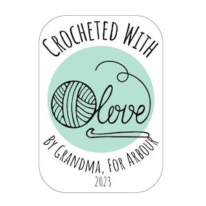 May include: A white label with a green circle in the centre. The circle contains a ball of yarn and a cursive word "love". The text on the label reads "Crocheted with love by Grandma, for Arbour 2023".