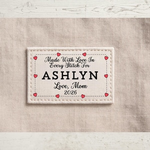 May include: A rectangular fabric label with the text "Made With Love In Every Stitch For ASHLYN Love, Mom 2026". The label is bordered with a dotted line and small red hearts. The background is a light beige fabric.