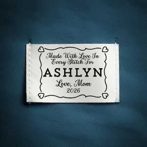 Puede incluir: Una etiqueta de tela blanca con texto negro y un borde decorativo. El texto dice "Made With Love In Every Stitch For ASHLYN Love, Mom 2026". La etiqueta está adherida a una tela azul oscuro texturizada.