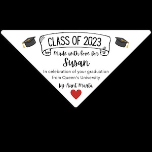 Op de afbeelding: Witte driehoekige banner met zwarte tekst die "CLASS OF 2023" luidt met een scrollontwerp en de woorden "made with love for Susan" eronder. De banner heeft ook twee zwarte afstudeerhoeden met gouden kwastjes aan elke kant. Onder de tekst staat "In celebration of your graduation from Queen's University by Aunt Marta" met een rood hart.