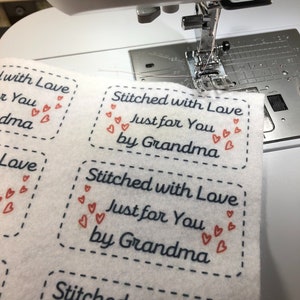 Könnte beinhalten: Nahaufnahme einer Nähmaschine, die ein weißes Stofflabel mit dem Text "Stitched with Love Just for You by Grandma" in blauer und roter Schrift näht. Das Label ist mit kleinen roten Herzen verziert.
