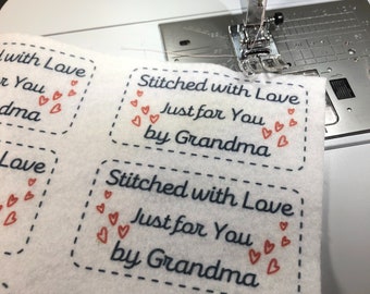 Puede incluir: Primer plano de una máquina de coser cosiendo una etiqueta de tela blanca con el texto "Stitched with Love Just for You by Grandma" en letras azules y rojas. La etiqueta está decorada con pequeños corazones rojos.