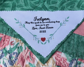 May include: A white triangle-shaped fabric label with green leafy vines and a red heart. The label has the text "Frelynn, May this quilt be an everlasting hug from me to you Love, Aunt Karen 2022".
